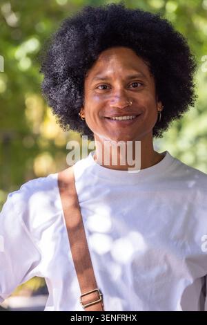 Mann lächelt, steht draußen im Park, trägt weißes T-Shirt, braunes Crossbody-Band, Nasenreif Stockfoto