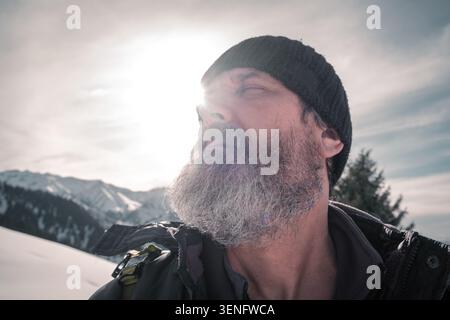 Nahporträt eines bärtigen, Reifen Mannes, der draußen vor schneebedeckten Bergen und Sonne blickt. Kommerzielles Stockbild für Reisen, Lifestyle, wi Stockfoto