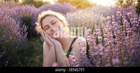 Eine junge Frau mit braunen Haaren lächelt sanft, während sie ihren Kopf auf den Händen legt, umgeben von Lavendelblüten in einem sonnendurchfluteten Feld Stockfoto
