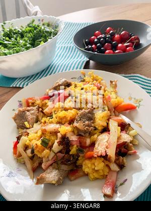 Eine Schüssel Gemüseeintopf mit Reis und Rindfleisch wird zusammen mit einem frischen Salat und eine Schüssel Beeren auf einem Holztisch in einer hellen Küche serviert Stockfoto