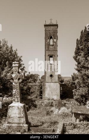 Schwarz-weiß-Foto eines keltischen Kreuzes auf dem Friedhof Ballinrobe, Irland, mit einem alten verlassenen Kirchturm und historischen Gräbern im Hintergrund Stockfoto
