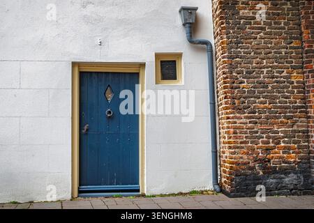 Eine dunkelblaue Tür mit einem diamantförmigen Fenster und einem Klopfer ist in eine weiße Stuckwand eingefasst, die an eine Ziegelstruktur angrenzt. Stockfoto