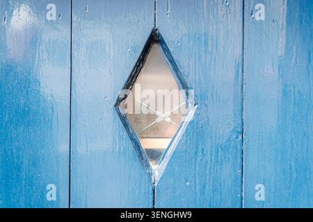 Eine diamantförmige Fensterscheibe in einer verwitterten blauen Holztür bietet einen Einblick in das Innere. Stockfoto