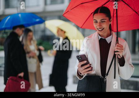 Junge Geschäftsfrau unter rotem Schirm lächelt am Telefon, während Kollegen im Regen plaudern Stockfoto