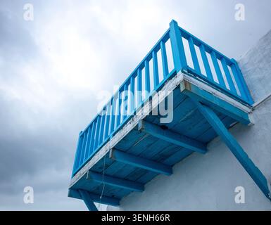 Flacher Blick auf einen traditionellen kykladischen Holzbalkon in hellem Blau in einem weißen Gebäude vor einem bewölkten Himmel. Stockfoto