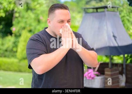 Junger Mann, der während der Allergiesaison im Freien in ein Gewebe niest. Authentisches Gesundheits- und Krankheitskonzept, das Erkältungssymptome, Grippe, Heuschnupfen, Stockfoto
