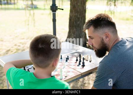 Ein bärtiger Mann und ein Junge spielen Schach an einem Parktisch unter einem Baum. Sie konzentrieren sich auf das Spiel und genießen eine gute gemeinsame Zeit. Stockfoto