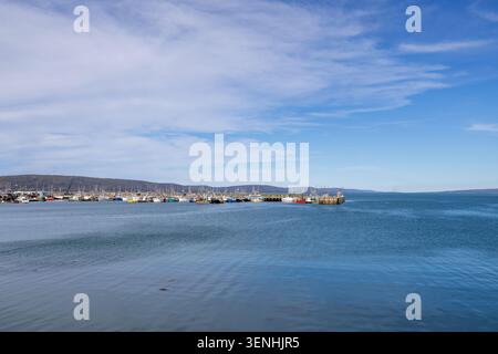 Breiter Panoramablick auf einen belebten Fischerhafen und ruhige Küstengewässer mit farbenfrohen Booten, die entlang der Uferpromenade unter dramatischen Wolken und blu liegen Stockfoto