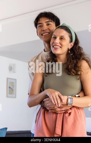 Paare umarmen und lächeln, während sie im Wohnzimmer mit olivfarbener Oberseite, Smartwatch und gerahmtem Druck stehen Stockfoto