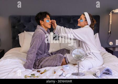 Asiatischer Mann und Afroamerikanerin, die auf dem Bett sitzen und blaue Flecken unter den Augen tragen, tragen Bademäntel Stockfoto