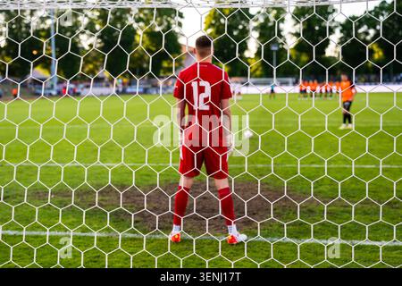 Sehen Sie durch das Netz eines Fußballtorhüters in einem roten Trikot, der sich darauf vorbereitet, während eines Fußballspiels einen Elfmeterschießen zu verteidigen. Stockfoto