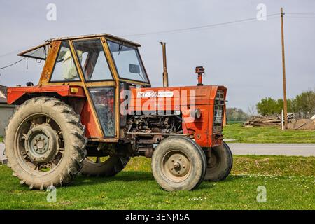 Vinkovci, Kroatien - 10. April 2026: Ein roter Vintage-Traktor, der auf einem grasbewachsenen Feld neben einem ländlichen Bauernhaus geparkt wurde und abgenutzte Reifen, freiliegender Motor und m zeigt Stockfoto