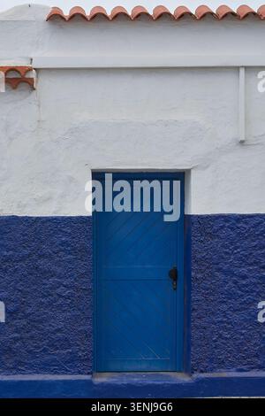 Vertikaler Blick auf eine traditionelle hellblaue Holztür auf einer rustikalen weiß-blau lackierten Stuckfassade mit Terrakotta-Dachziegeln. Stockfoto