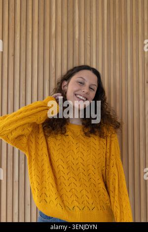 Frau in 20ern stehend, lächelnd, berührend den Hals vor Holzlamellenwand, in Senfpullover, Ohrringen Stockfoto