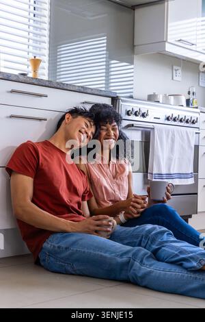 Verschiedene Paare sitzen auf gefliestem Küchenboden, lehnen sich gegen den Ofen und halten Keramikbecher Stockfoto