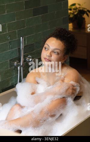 Afroamerikanische Frau sitzt in freistehender Wanne vor dunkelgrünen Fliesen, bedeckt mit weißem Schaum Stockfoto