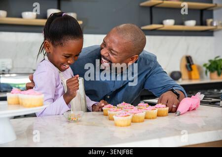 Afroamerikanischer Vater führt Mädchen in Schürze, die Cupcakes auf der Theke mit Glasur und Streuseln dekoriert Stockfoto