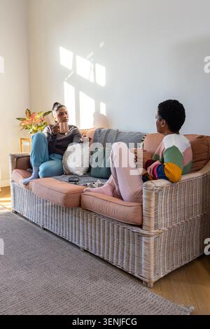 Verschiedene Freundinnen schlürfen an Keramikbechern und plaudern auf dem Rattansofa im Wohnzimmer Stockfoto