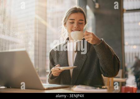 Glückliche junge Erwachsene kaukasische Frau, die warmen Kaffee in einem modernen Café in der Nähe des Fensters trinkt Stockfoto