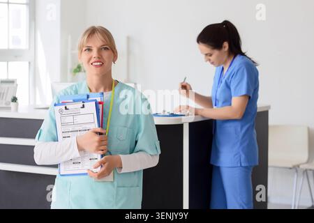 Lächelnde Krankenschwester mit medizinischen Karten und Klemmbrett im Krankenhausaufnahmebereich, eine andere medizinische Fachkraft arbeitet am Schalter im Rücken Stockfoto
