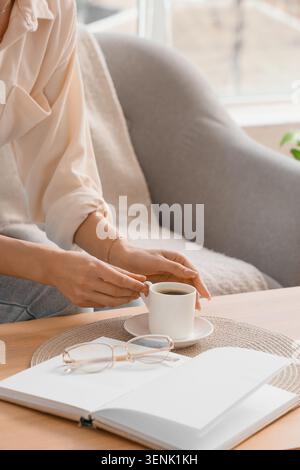 Frauenhände halten eine Kaffeetasse auf einem Holztisch mit offenem Notizbuch und Brille, gemütliche Morgenroutine, Frauenhände halten sanft einen weißen Kaffee Stockfoto