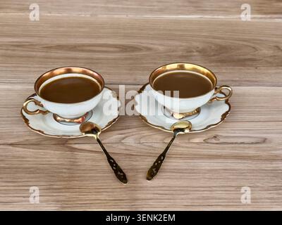 Zwei Tassen Kaffee mit Milch in antiken Kaffeetassen mit silbergoldenen Löffeln auf Untertassen auf einem Eichentisch. Nahaufnahme von vorne. Stockfoto