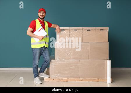 Lächelnder Liefermann in Sicherheitsweste mit Klemmbrett, der auf gestapelten Kartonschachteln für Versand und Logistik gebeugt ist, die Kartons sind versandfertig und Stockfoto