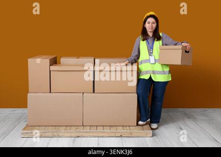 Weibliche Lagerarbeiterin in Sicherheitsweste und Schutzhelm mit Pappkarton, Logistik und Lieferservice, mehrere gestapelte Pappkartons auf einem Holz Stockfoto