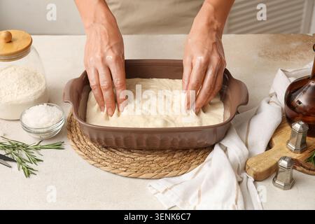 Frauenhände, die Focaccia-Teig in der Backform mit Mehl, Salz und Rosmarin auf der Küchenzeile drücken Stockfoto
