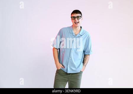 Glücklicher junger Mann in blauem Hemd und grüner Hose lacht mit Händen in Taschen, lässiges Modeporträt, er trägt eine Brille und hat Tätowierungen sichtbar auf seinem Stockfoto