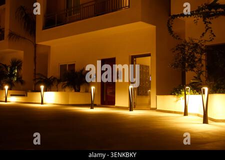 Moderne Wohngebäude Eingang bei Nacht mit warmer Außenbeleuchtung und Palmen, Palmen und üppiger Landschaftsgestaltung ergänzen das Außendesign Stockfoto