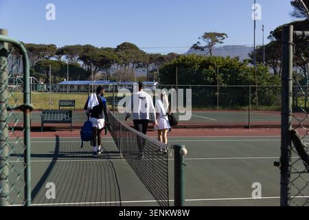 Verschiedene Tennismannschaftskameraden laufen entlang der Mittellinie des Platzes, vorbei am Netz auf einem umzäunten Platz mit Taschen Stockfoto