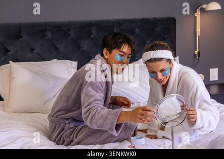 Verschiedene Paare, die auf dem Bett sitzen und weiche Bademäntel tragen und einen runden Spiegel halten, unter den Augen Stockfoto