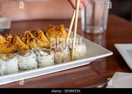 Eine detaillierte Nahaufnahme, die den Moment feststellt, in dem eine Person mit Holzstäbchen eine Sushi-Rolle mit Sesamkruste von einer weißen Platte aufnimmt. Die Platte f Stockfoto