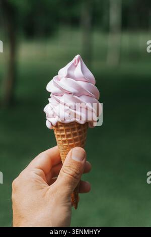 Die Hand hält einen Wirbel rosafarbener Softeis in einer Waffelhülle vor einem üppigen, sonnendurchfluteten grünen Hintergrund, der perfekt das Wesen eines verkörpert Stockfoto