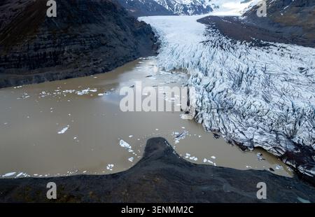 Der Svinafellsjokull-Gletscher bricht auseinander, während er zu einem schlammigen Wasserkörper schmilzt. Eisberge schweben im Wasser, während felsiges Land die Szene umgibt. Der Bereich Stockfoto