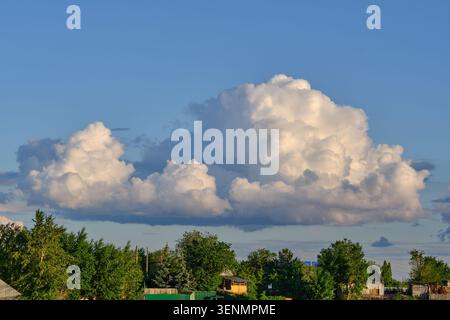 Riesige weiße Wolken schweben am sonnigen Sommertag über grünen Dorfbäumen vor klarem blauem Himmel Stockfoto