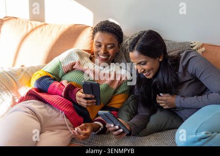 Verschiedene Freundinnen lachen und teilen Smartphones auf dem Sofa mit gemustertem Überwurf und Kissen Stockfoto