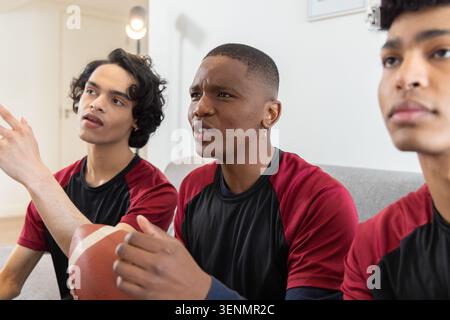 Verschiedene männliche Teenager-Freunde in passenden Trikots sitzen zu Hause auf grauem Sofa und halten Fußball Stockfoto