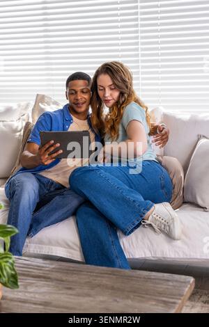 Verschiedene Paare sitzen dicht auf hellem Sofa, teilen sich ein Tablet und lehnen sich ineinander Stockfoto
