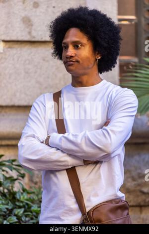 Afroamerikaner, der mit gekreuzten Armen vor einer Steinmauer steht und braune Umhängetasche trägt Stockfoto