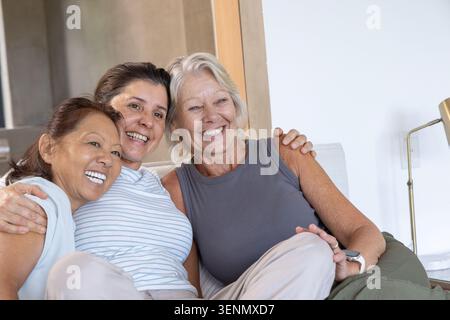 Verschiedene Freundinnen, darunter Senioren, die sich zu Hause auf einem leichten Sofa umarmen und mit Smartwatch lächeln Stockfoto