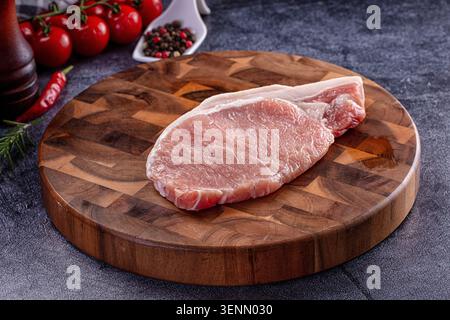 Rohes, frisches Schweinefleischfilet-Steak auf rundem Holzschneidebrett, begleitet von Kirschtomaten, Chili-Pfeffer, Pfeffer und Rosmarin auf dunklem Hintergrund Stockfoto