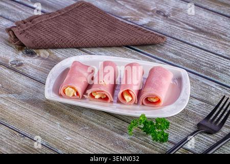 Vier mit Käse gefüllte, gerollte Schinkenscheiben auf einem weißen rechteckigen Teller, auf einem rustikalen Holztisch mit einer braunen Serviette mit Waffelstruktur gestellt Stockfoto