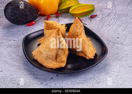 Drei knusprige gebratene Samosas auf einem schwarzen Teller. Dieser beliebte indische Snack ist umgeben von frischen Zutaten wie Paprika, Sternfrüchten und Chili, cre Stockfoto