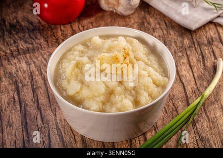 Ein Nahaufnahme einer warmen Schüssel mit cremigem Blumenkohlpüree, garniert mit Paprika. Das Gericht wird auf einem rustikalen braunen Holztisch mit frischen Zutaten präsentiert Stockfoto