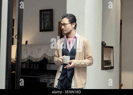 Junger asiatischer Mann genießt eine Tasse Kaffee, während er sich drinnen entspannt Stockfoto