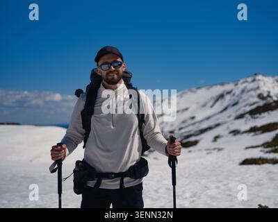 Ein glücklicher Wanderer trägt Jacke, Baseballmütze und Sonnenbrille und hält Wanderstöcke. Schneebedeckte Berghänge und klarer blauer Himmel im Frühling Karpaten Mo Stockfoto