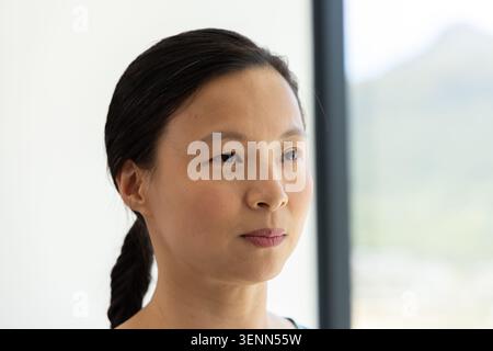 Asiatische Frau mittleren Alters, die in der Nähe eines hohen vertikalen Fensters steht und geflochtene Haare und dünne Riemen zeigt Stockfoto