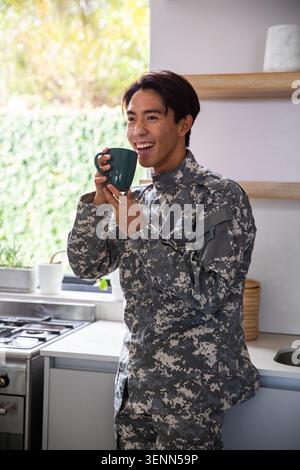 Asiatischer Mann, der an der Theke in der hellen Küche steht, trägt eine Tarnjacke, die einen grünen Becher lächelnd hält Stockfoto